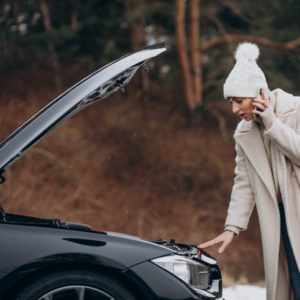 A woman in winter clothing looks under the hood of a car on the side of the road.